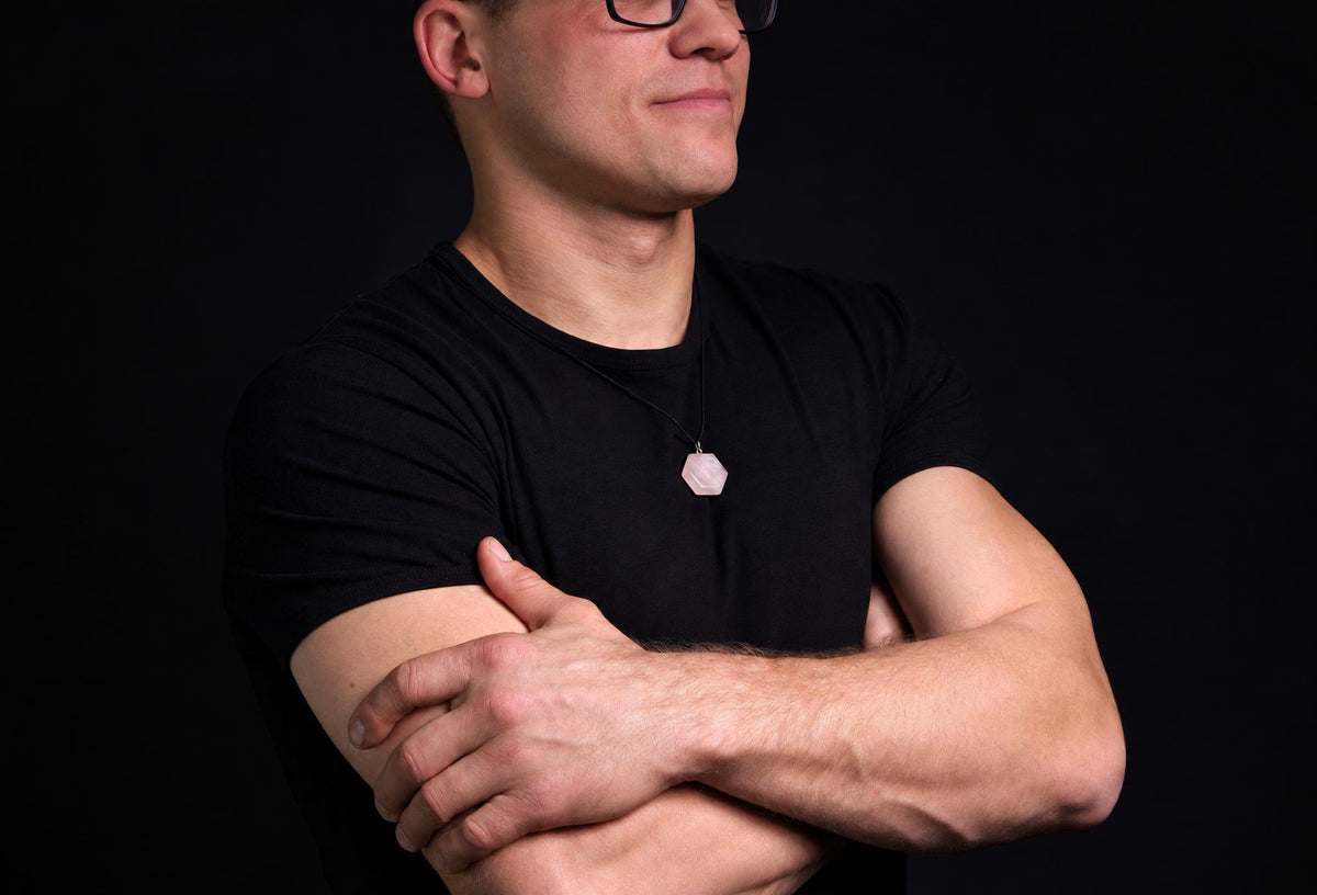 Vyras juodu marškinėliu dėvi šviesiai rožinį šešiakampį pakabuką ant kaklo, stovėdamas su kryžtomis rankomis prieš tamsų foną.