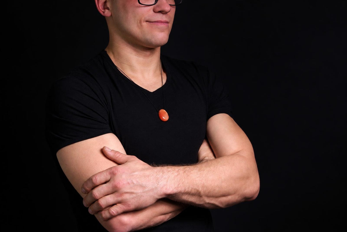 Vyras juodu marškinėliu dėvi pakabuką su oranžiniu, ovaliu karneolio akmeniu ant tamsaus fono.