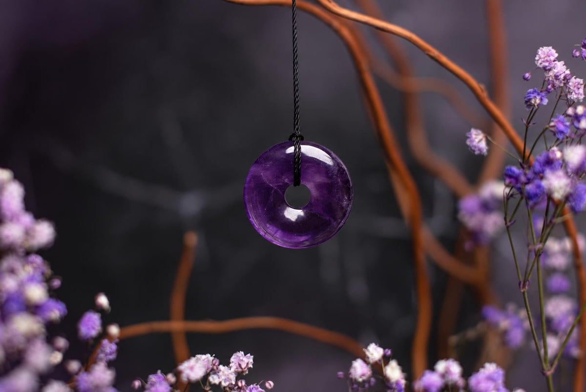 Violetinis disko formos pakabukas su apvalia anga centre, kabantis ant juodos virvelės tarp smulkių violetinių žiedų ir rudų šakų tamsiame fone.