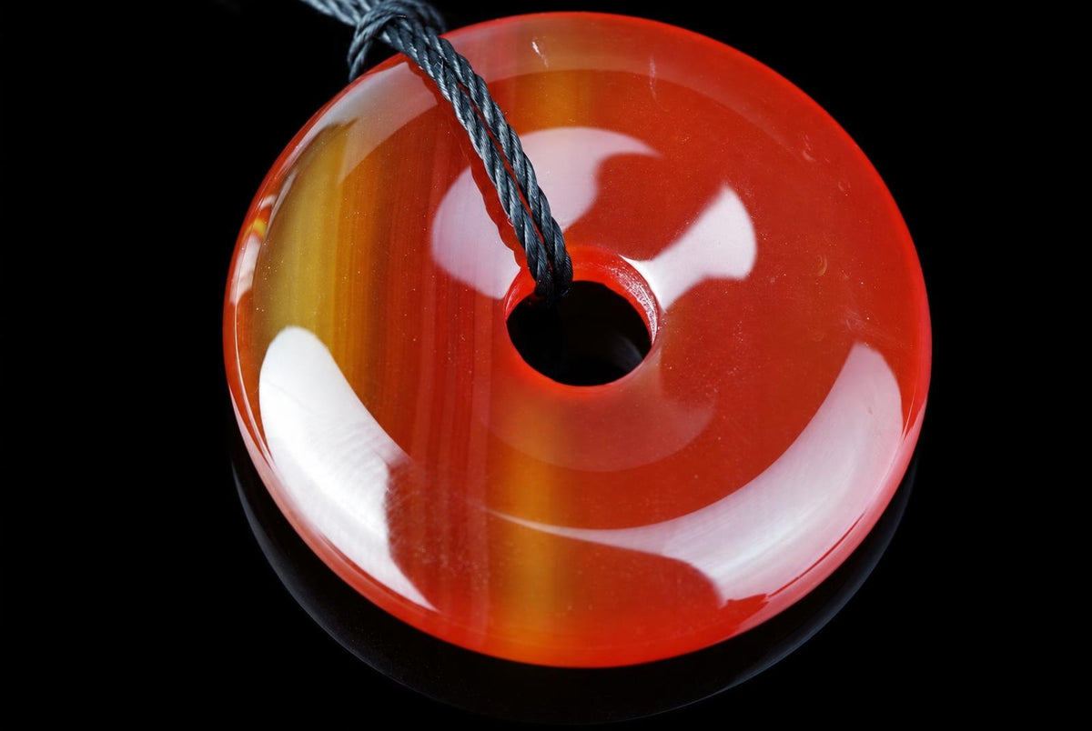 Blizgus apvalus raudonai oranžinis pakabukas su gelsvomis juostomis ir skyle centre, per kurią perverta tamsi pinta virvelė; stambus planas juodame fone.
