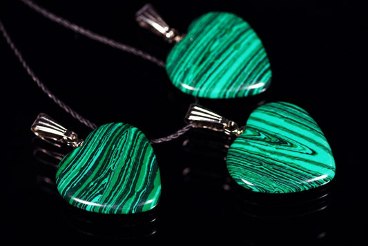 Trys širdies formos pakabukai su žalios ir tamsiai žalios spalvos juostuotais akmenimis ant tamsaus paviršiaus.