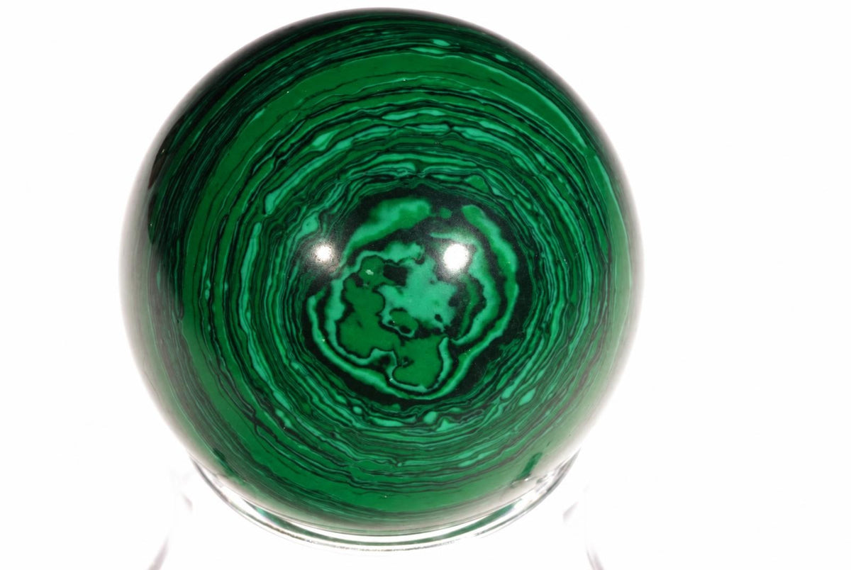 Žalia malachito sfera su juodomis spiralėmis, stovinti ant skaidraus pagrindo baltame fone.