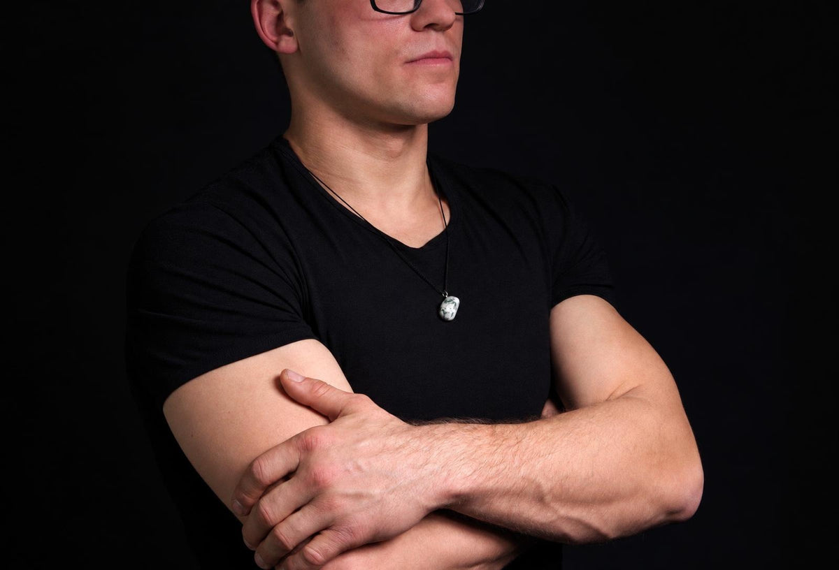 Vyras juodu marškinėliu dėvi kaklo apyrankę su baltos ir žalios spalvos akmenuko formos pakabuku ant tamsaus fono.