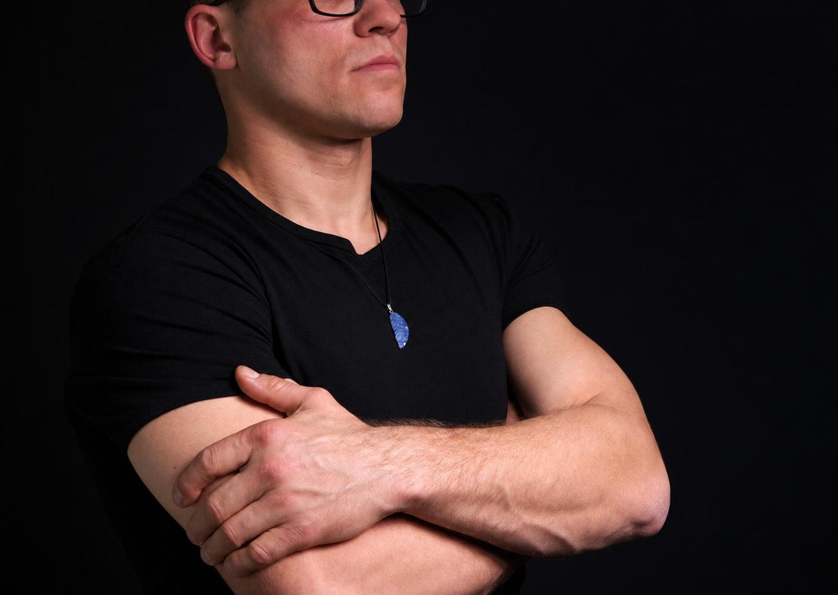 Vyras juodu marškinėliu dėvi kaklo apyrankę su mėlynos spalvos, sparnelio formos pakabuku ant tamsaus fono.