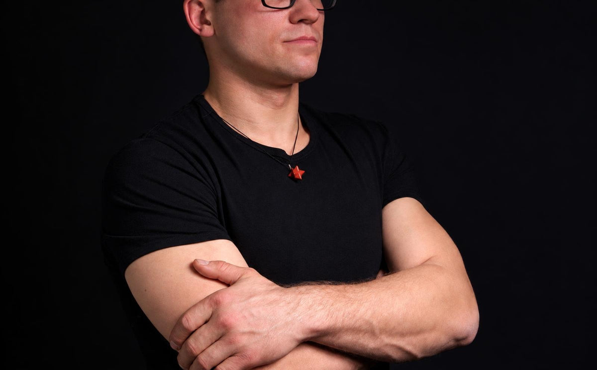 Vyras juodu marškinėliu dėvi raudoną žvaigždės formos pakabuką ant kaklo, stovėdamas su kryžtomis rankomis prieš tamsų foną.