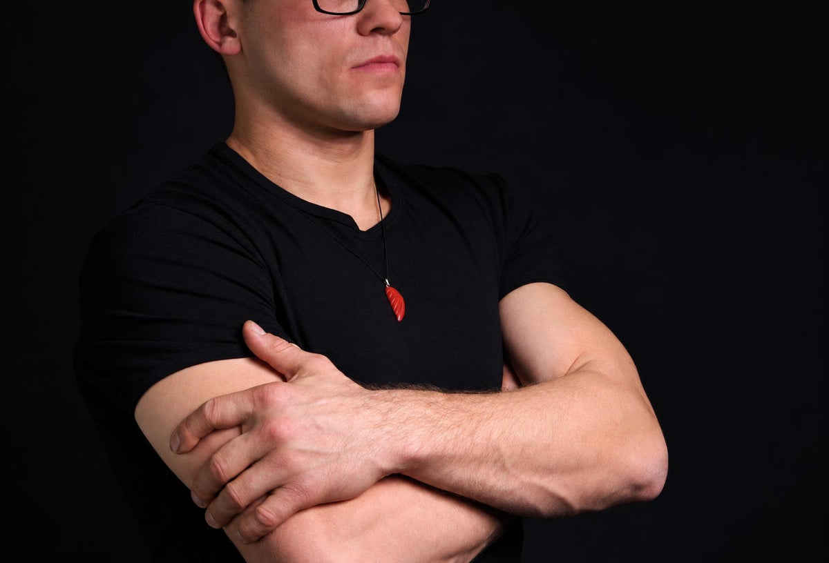 Vyras juodu marškinėliu dėvi raudoną, sparno formos pakabuką ant kaklo. Fonas tamsus.