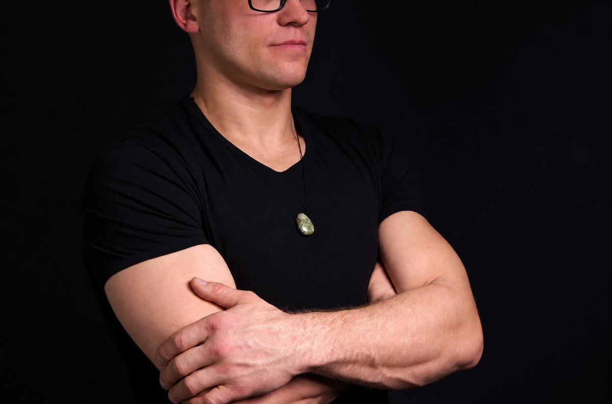 Vyras juodu marškinėliu dėvi pakabuką su žaliu, lašo formos akmeniu ant kaklo. Fonas tamsus.