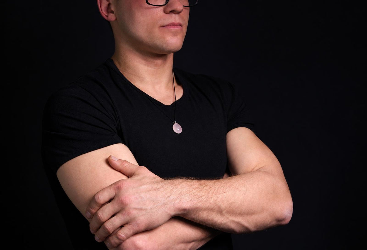 Vyras juodu marškinėliu dėvi kaklo apyrankę su rožinės spalvos, apvaliu pakabuku ant tamsaus fono.