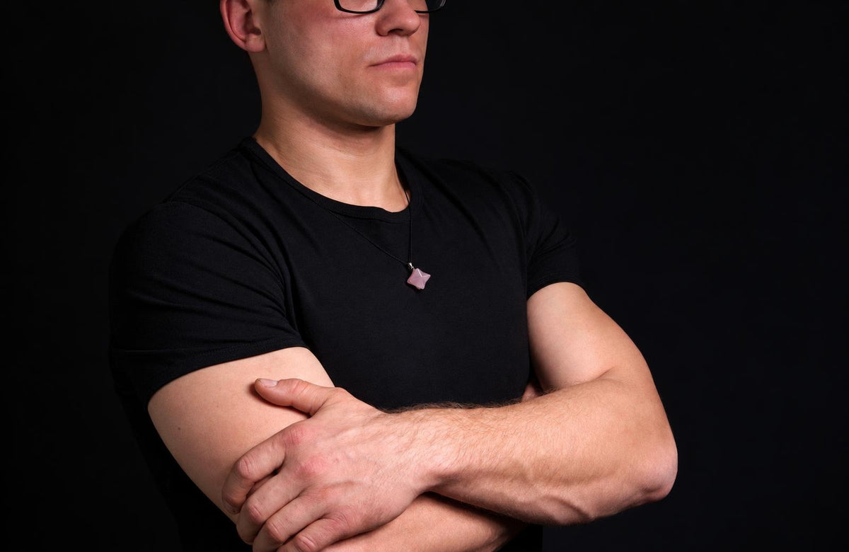 Vyras juodu marškinėliu dėvi kaklo apyrankę su rožinės spalvos, žvaigždės formos pakabuku ant tamsaus fono.