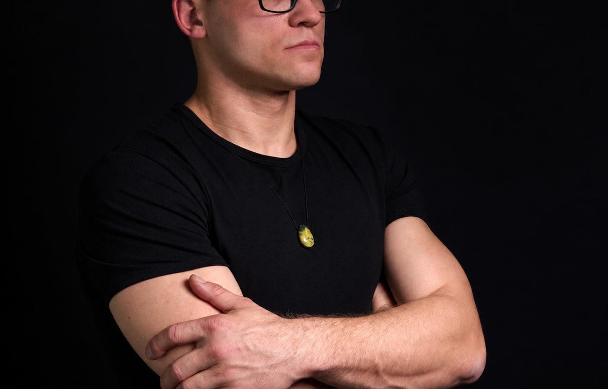 Vyras juodu marškinėliu dėvi kaklo pakabą su geltonu, lašo formos akmeniu ant tamsaus fono.