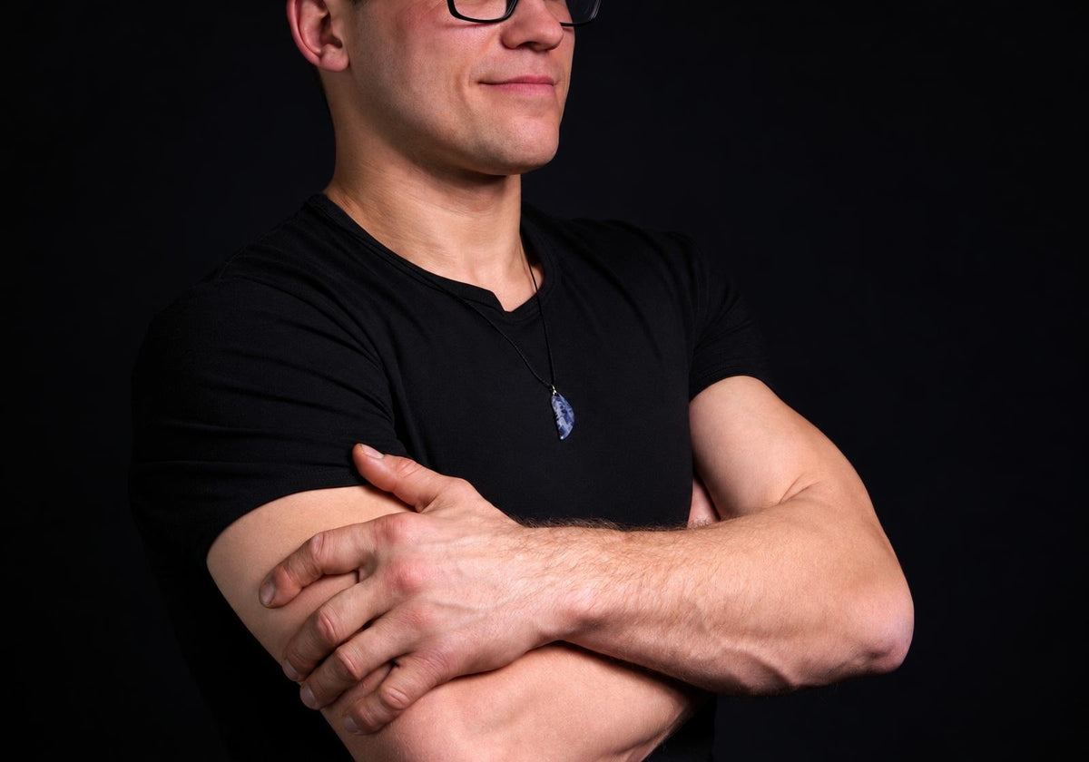 Vyras juodu marškinėliu dėvi pakabuką su mėlynai baltu akmeniu ant kaklo, stovėdamas prieš tamsų foną.