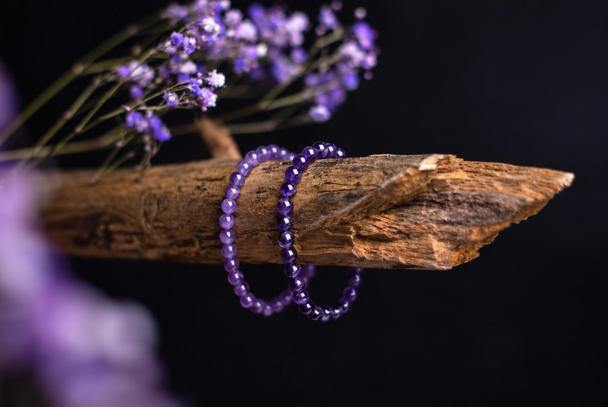 Dvi violetinės apyrankės su rutuliukais ant medinio šakos, fone – tamsus fonas ir violetiniai gėlės.