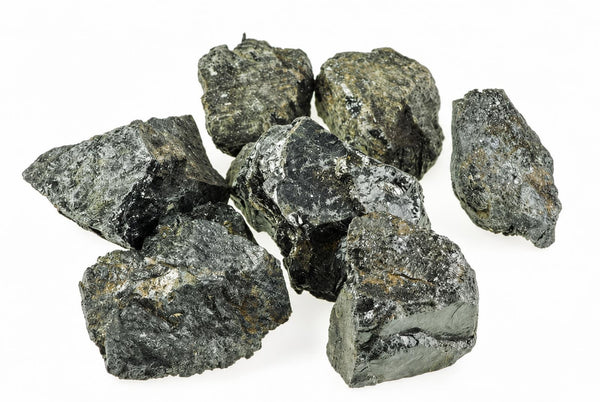 Keli neapdirbti tamsiai pilki ir juodi mineralų gabalai su šviesiais atspalviais ant baltos fono.