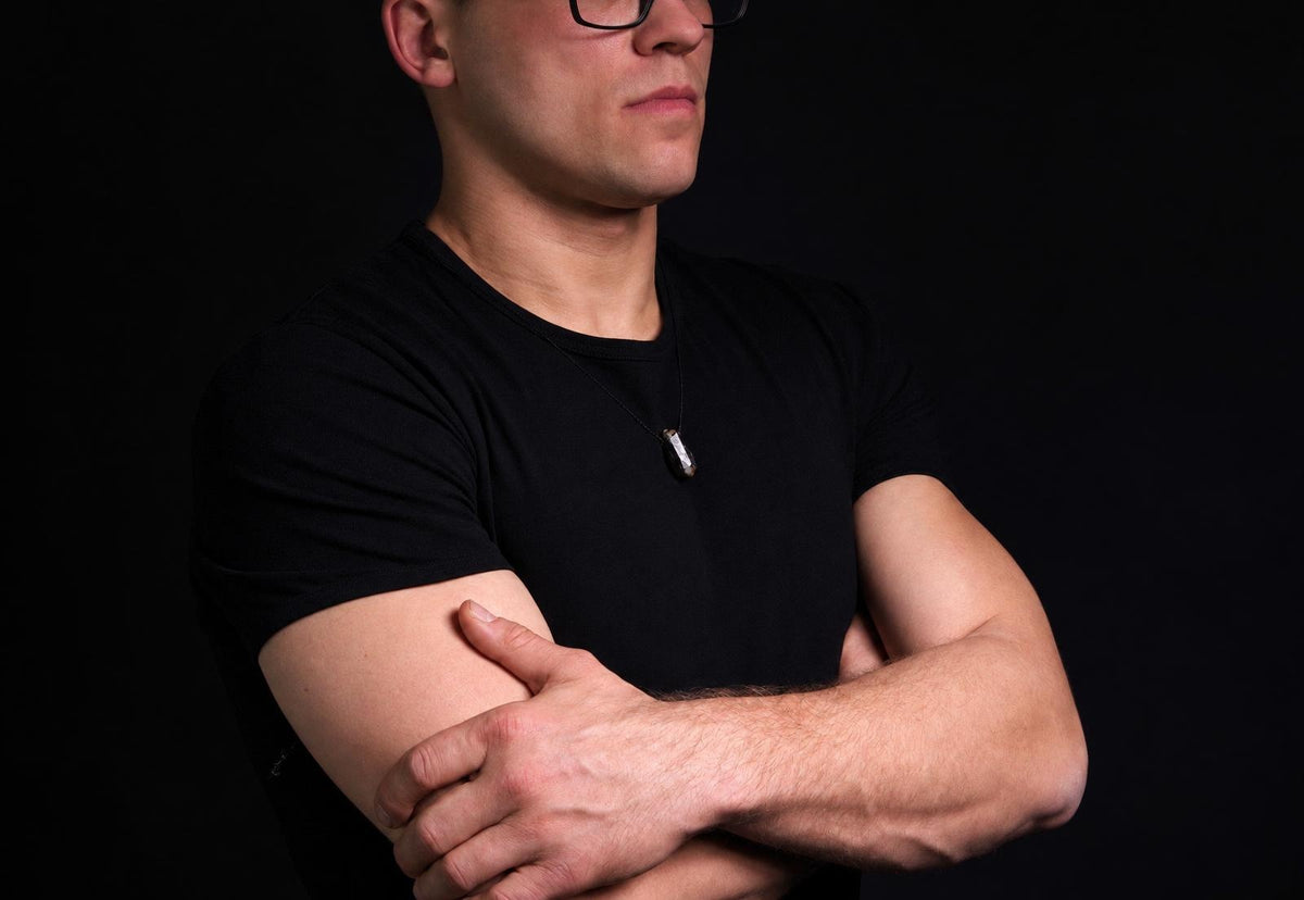 Vyras juodu marškinėliu dėvi pakabuką su tamsiai pilku, neapdorotu mineraliniu akmeniu ant kaklo.
