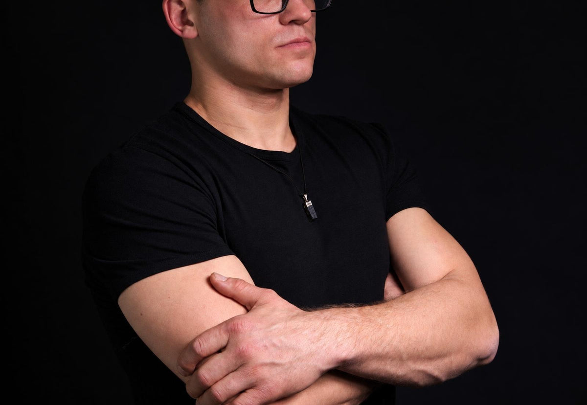 Vyras juodu marškinėliu su tamsiai pilku pakabuku ant kaklo, stovintis prieš tamsų foną.